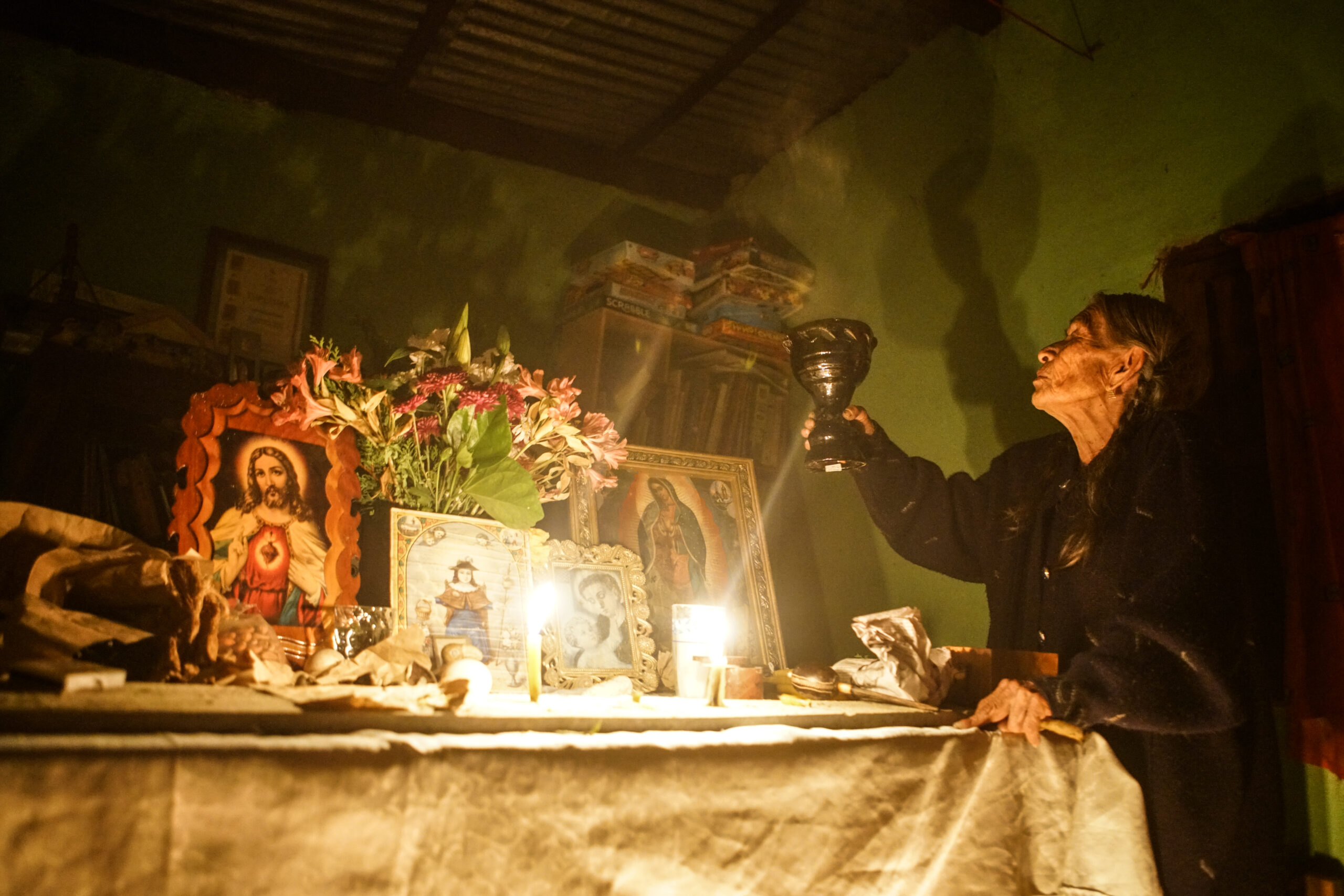 A Mazatec curandera in the middle of a ritual holding up a cup at an altar with catholic images and candles.