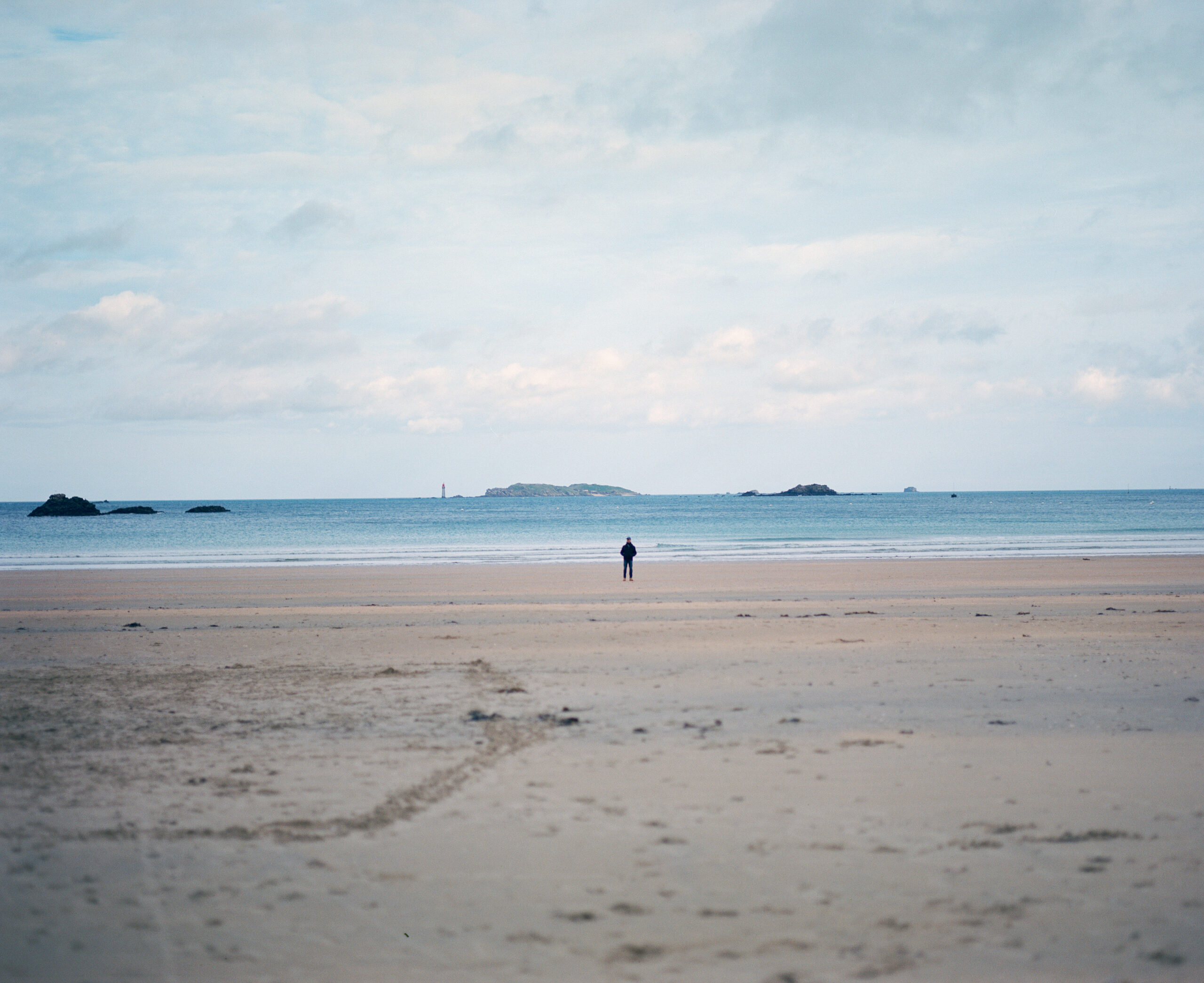 Luc Marescot stands far away from the camera, on the beach, in front of the sea, at Saint Lunaire in France.