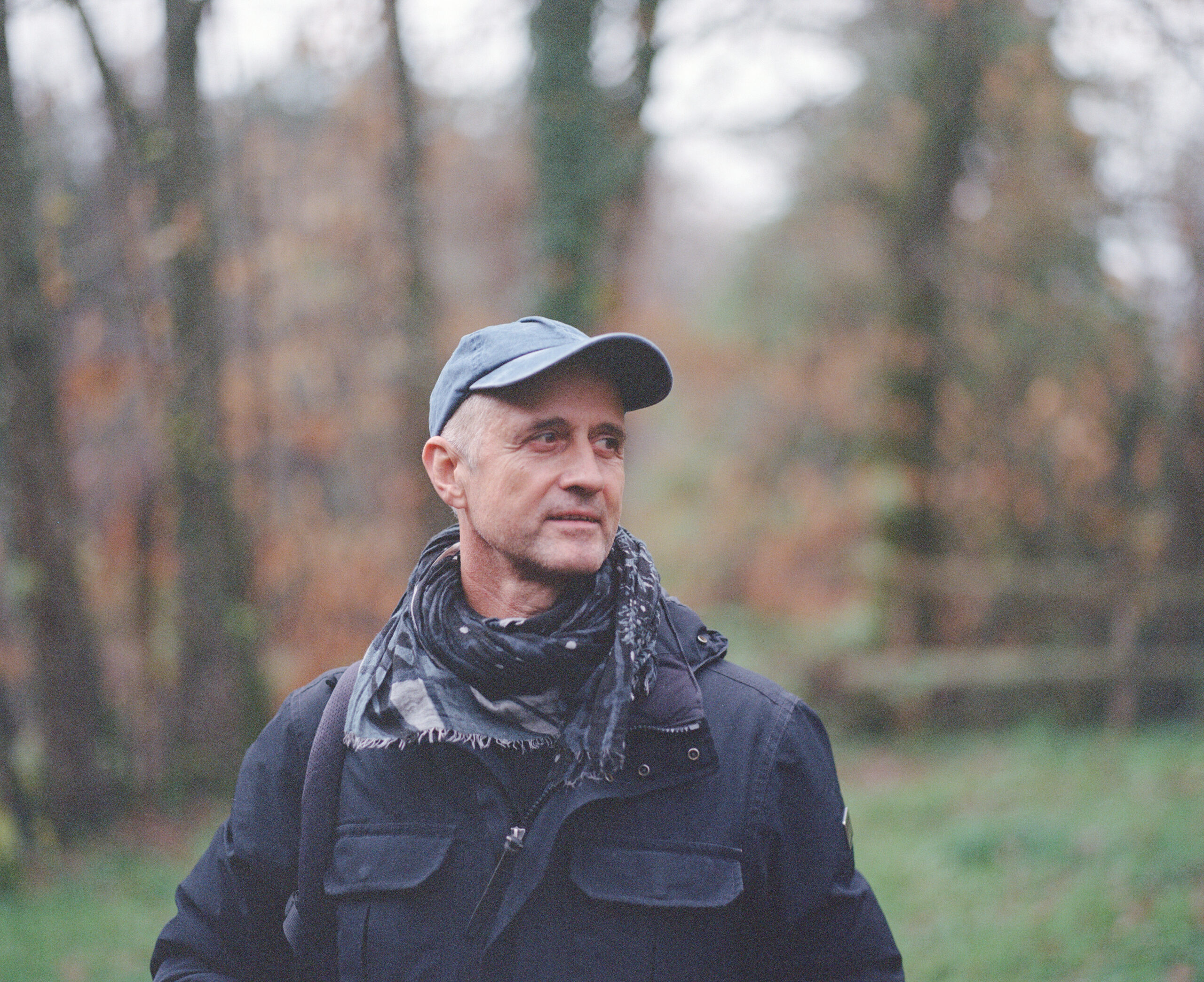 Luc Marescot is wearing a cap while standing in a garden, wearing clothes for what appears to be cold weather, looking away from the camera which reveals his profile.