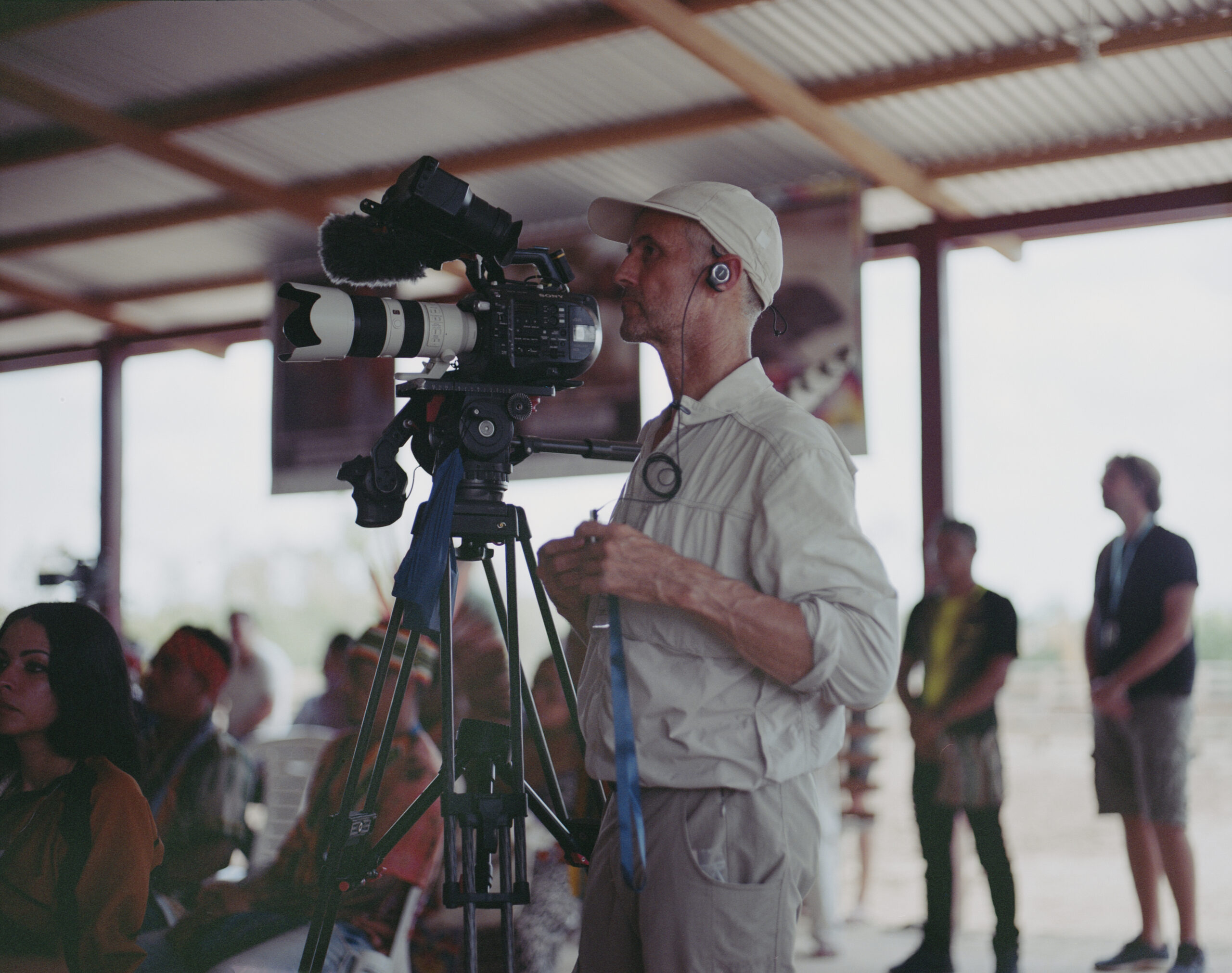 Luc Marescot stands behind a camera, with an earpiece in his left ear, filming amongst a crowd of people gathered for the IV Indigenous Ayahuasca Conference in Acré, Brazil.