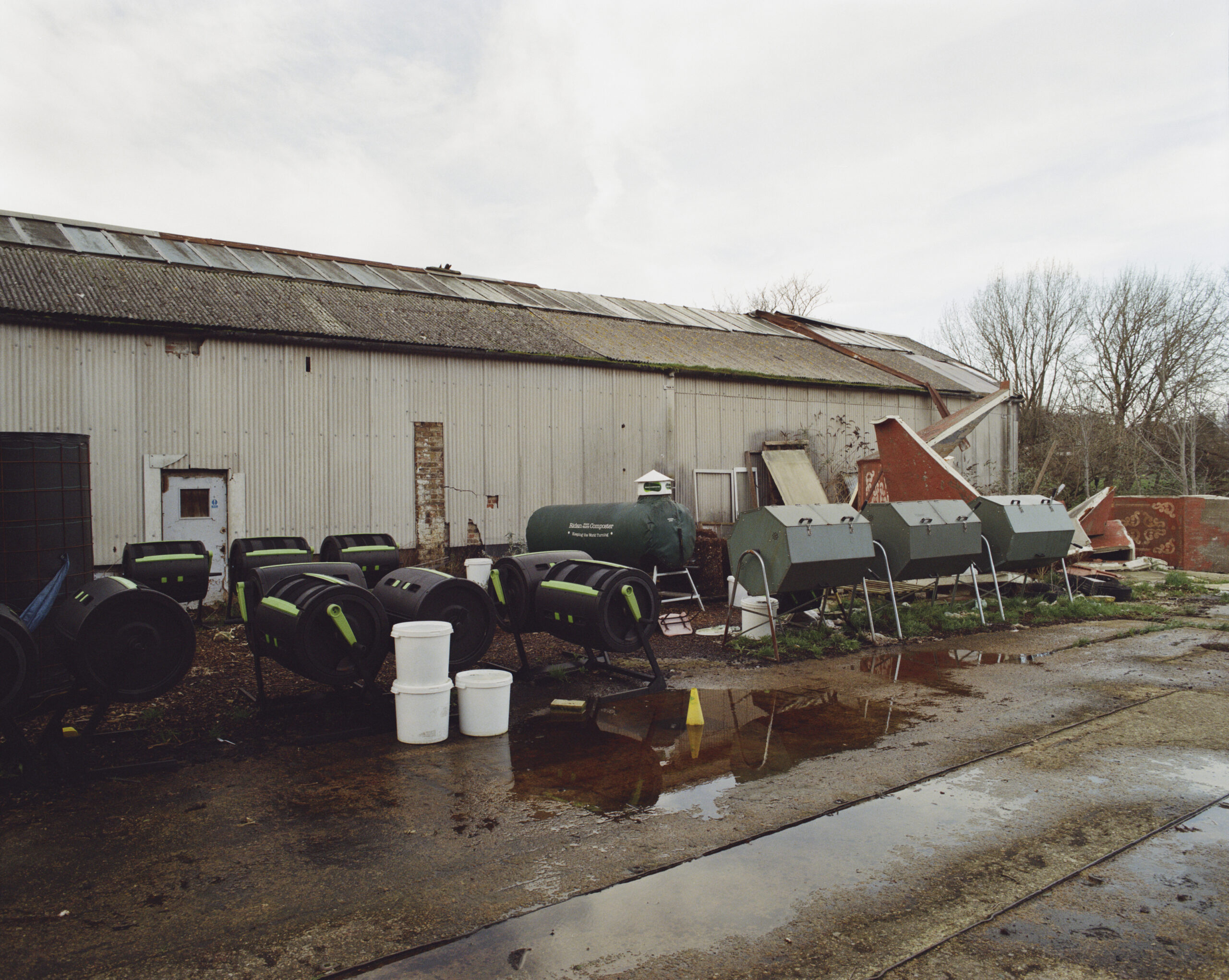 In front of a large shed are a group of tumblers that Michael uses to create compost. To the right are the bigger green tumblers, the smaller black tumblers are on the left. In front is a concrete road covered in puddles of water.