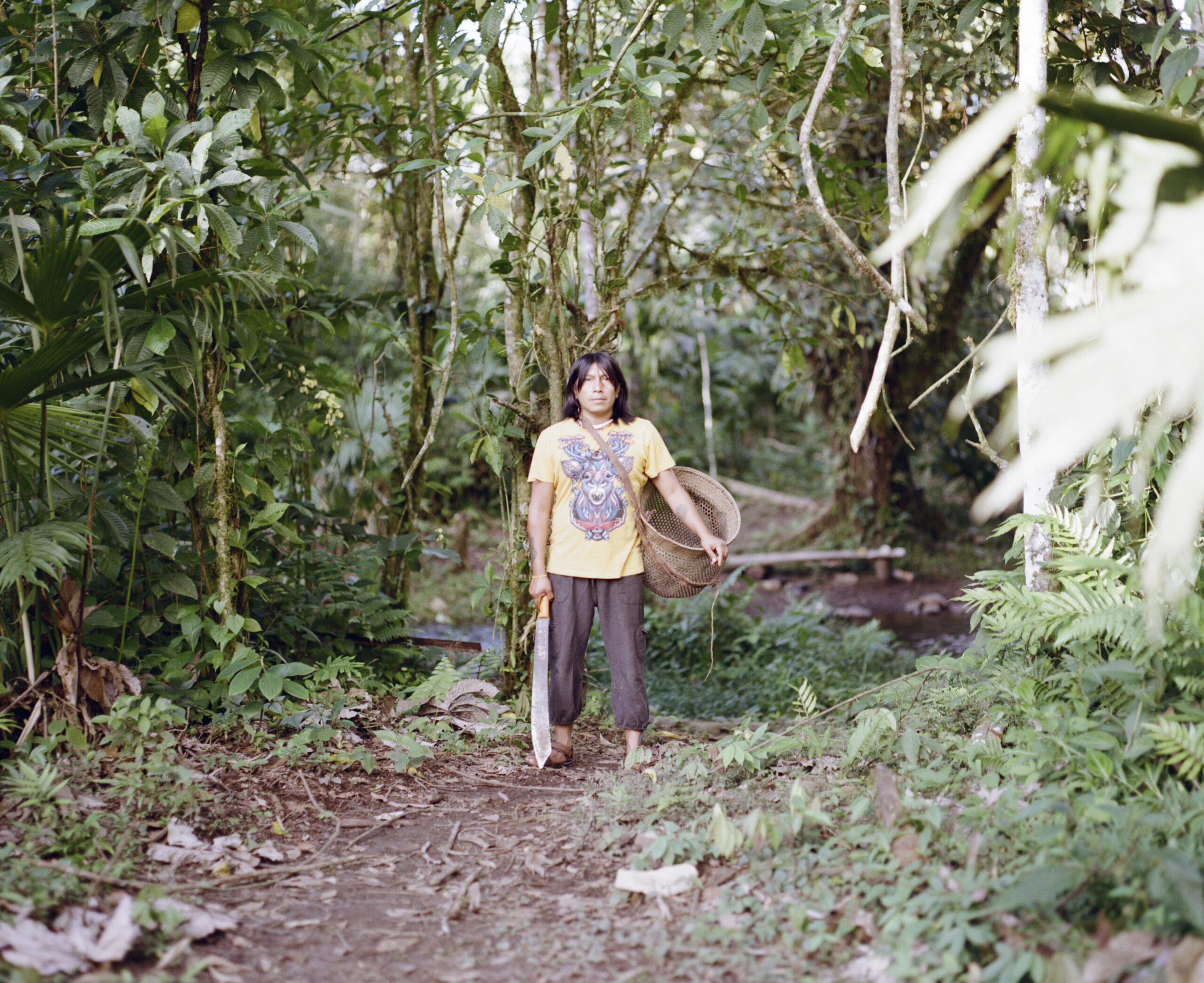 Yanda Montahuano Ushigua de pie a lo largo de un camino, sosteniendo una cesta y un machete, entre la selva en Itia Mamá Minga.