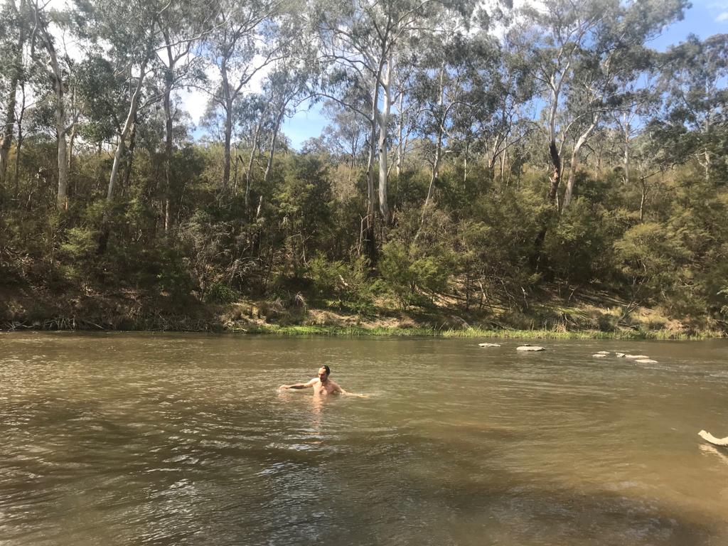 The author, Anton, swims in the middle of the brown River, with trees on the opposite bank, behind him.